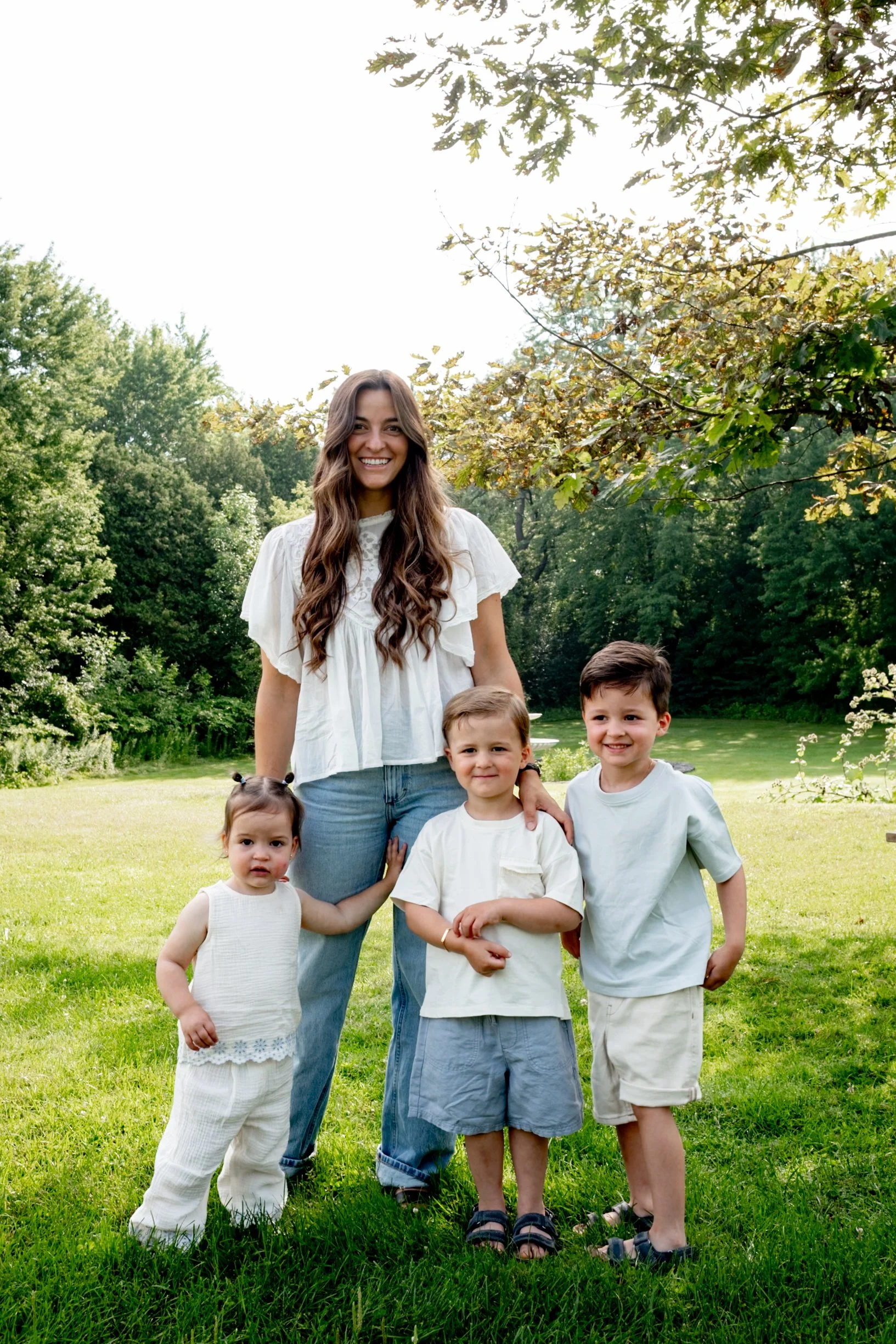 A woman and four children, standing on a green lawn in a park with trees behind them. The woman has long wavy hair and is smiling, wearing a white top and jeans. The children are dressed in light-colored clothing and are standing closely, interacting