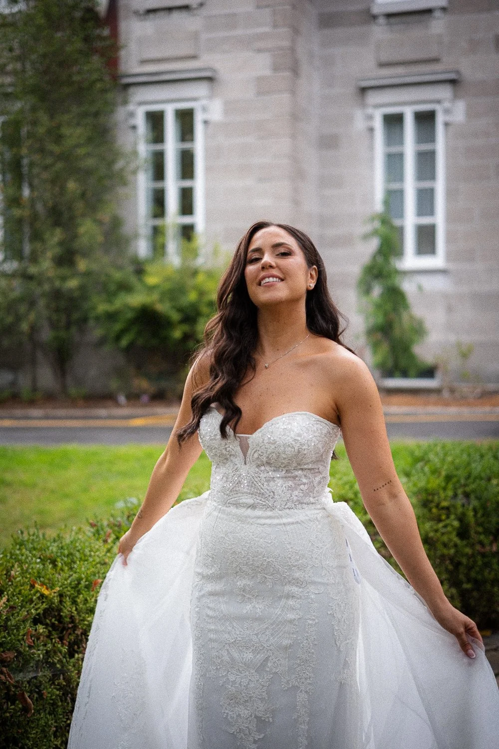 Montreal wedding photographer photographed a bride in a white wedding dress smiling outdoors in front of a stone building and greenery. 