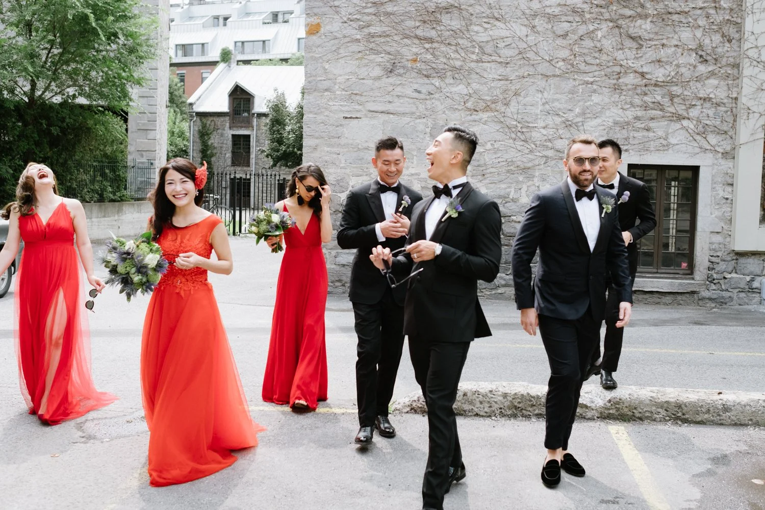 Group of six people dressed in formal attire, standing on a city street outside a stone building. Four women in red dresses are holding bouquets of flowers, and two men in black tuxedos with bow ties are smiling and walking.