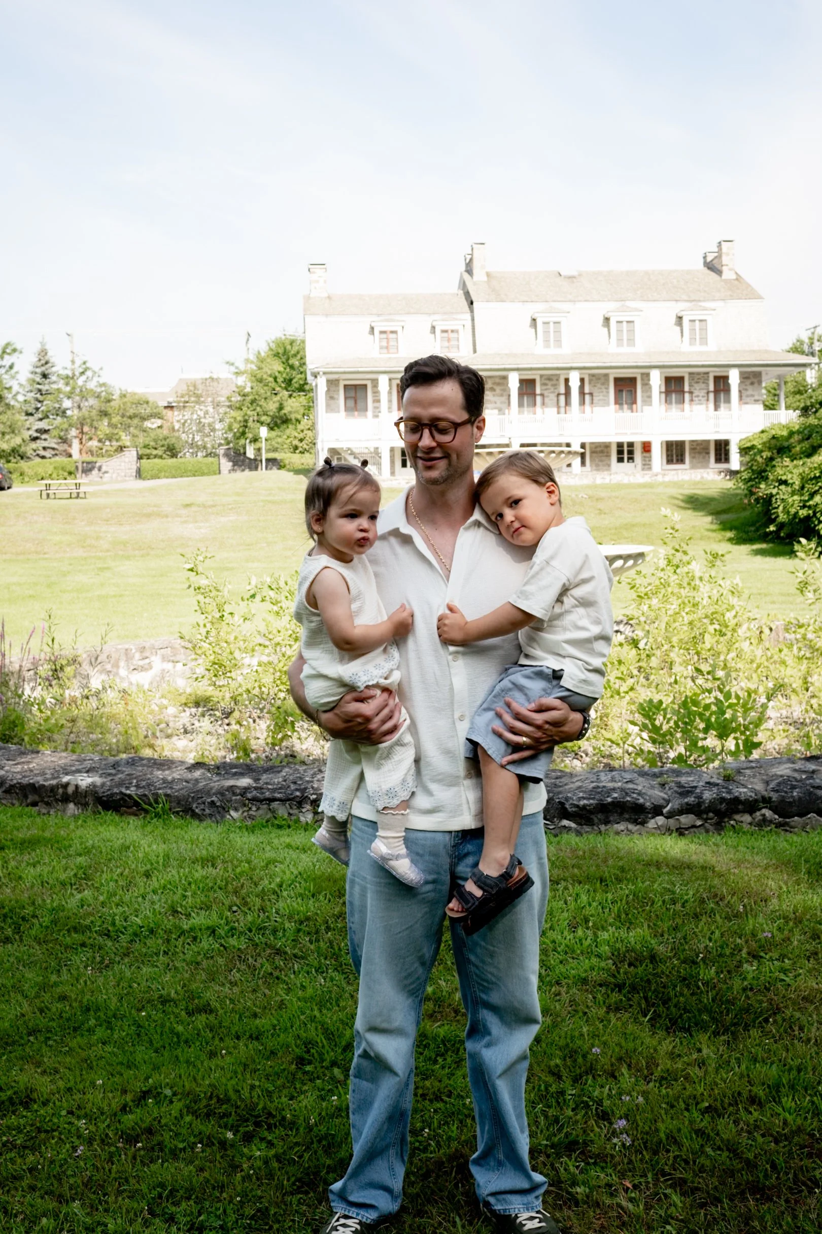 A man holding two children outdoors in a grassy area with a large house in the background. The man is wearing glasses and a light shirt, while the children are dressed in light clothing, one girl and one boy.