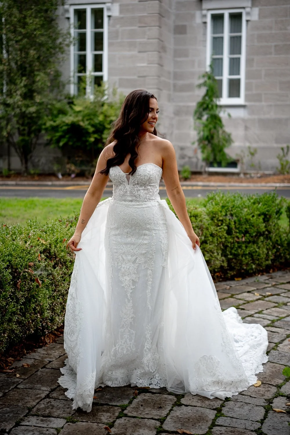 Montreal wedding photographer photographed a young bride in a white wedding dress stands outdoors on a cobblestone path, smiling and holding the sides of her dress. Behind her is a stone building with white-framed windows and greenery.