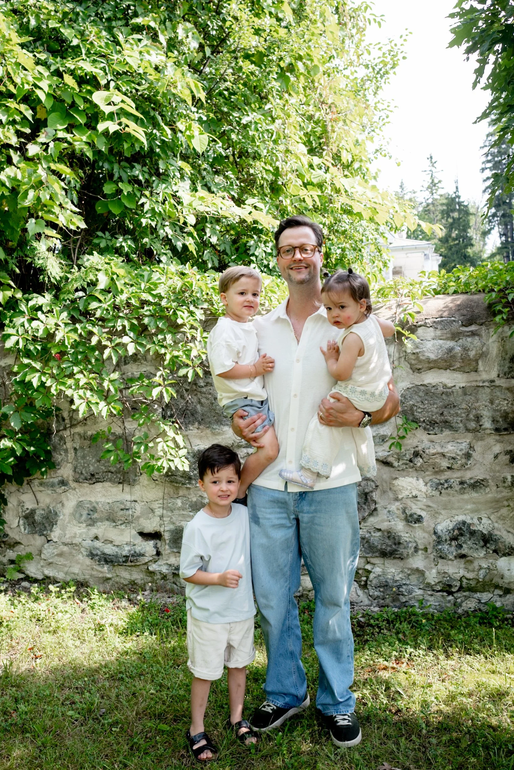 A man holding two young girls outside by a stone wall with lush green trees and bushes behind them. A boy stands in front of the man, all smiling and wearing casual light-colored clothing.