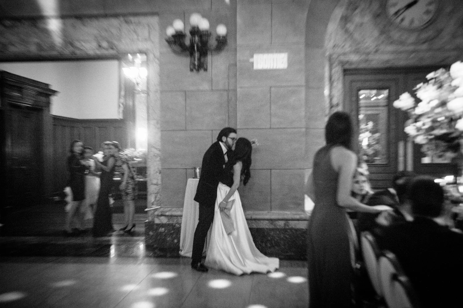 A black and white photo of a wedding reception with a bride and groom kissing in the center. The groom is wearing a black suit, and the bride is in a white wedding dress. Several guests are visible in the background, with some standing and others sit