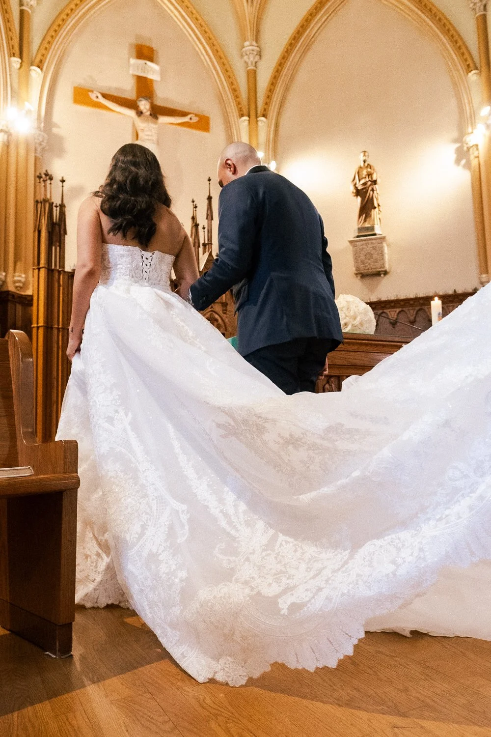 A couple getting married in a church, the bride in a white wedding gown with a long train, and the groom in a dark suit, standing before the altar with a crucifix and religious statues.