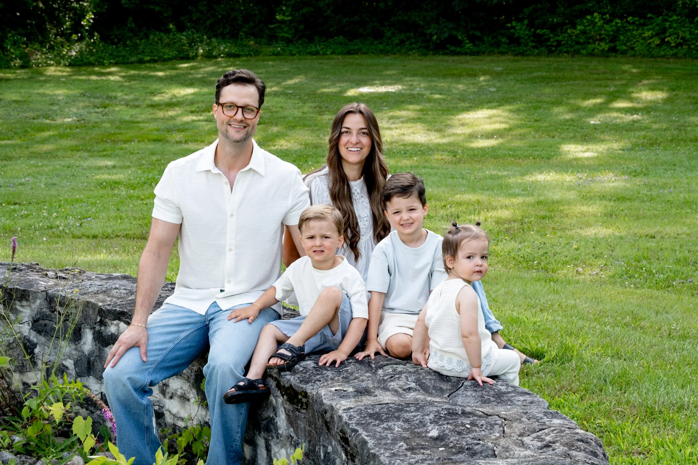 A family of five sitting outdoors on a rock in a grassy park, smiling for the camera, with trees in the background.