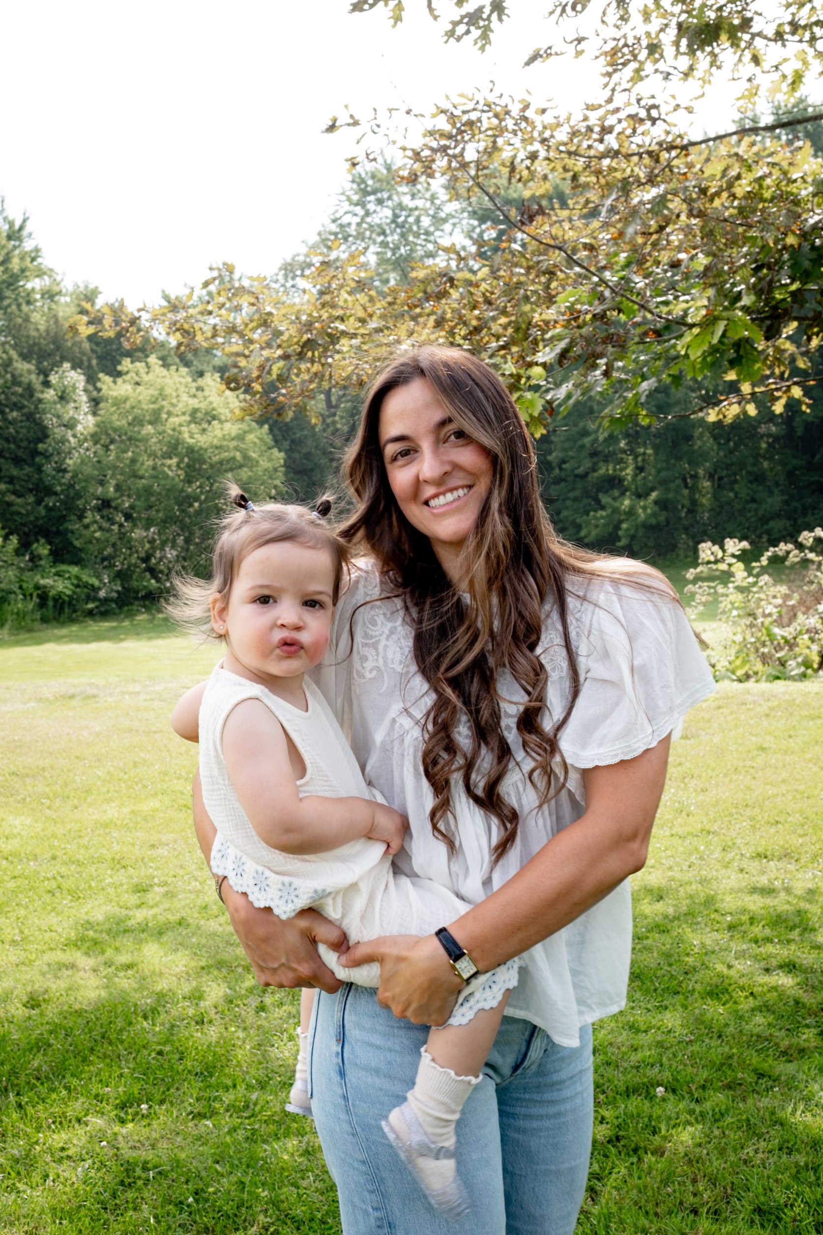 A woman with long brown hair smiling holds a young girl with light brown hair styled in pigtails, both standing outdoors on a sunny day with green trees and grass in the background.