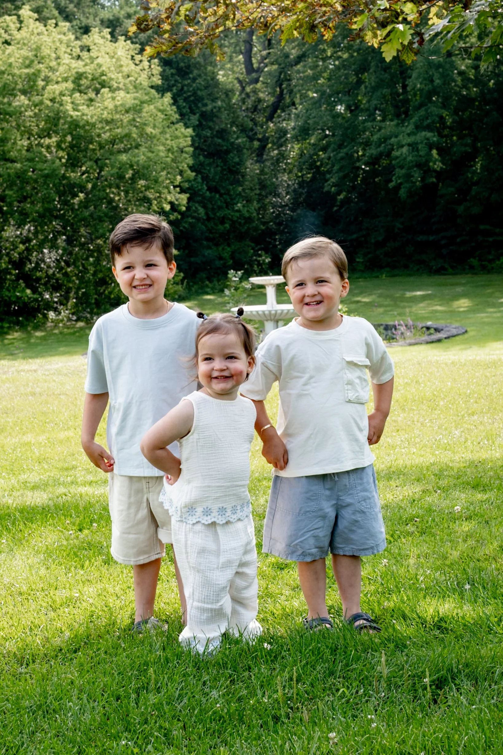 Three children standing on grass in a park, smiling, with trees and a fountain in the background during daytime.