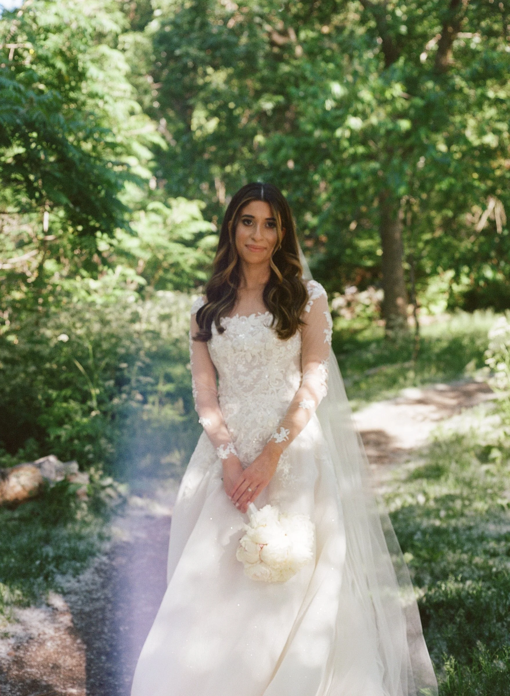 A bride in a lace wedding gown holding a bouquet, standing on a forest path surrounded by green trees.