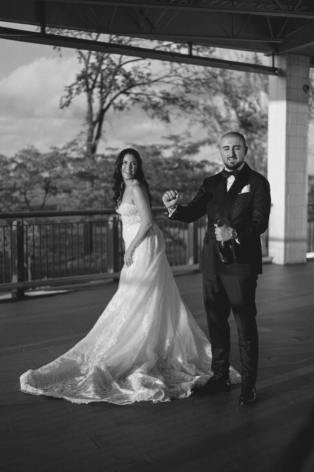 A black and white photo of a bride in a wedding gown smiling and standing with a groom in a tuxedo on a balcony, with trees and clouds in the background.