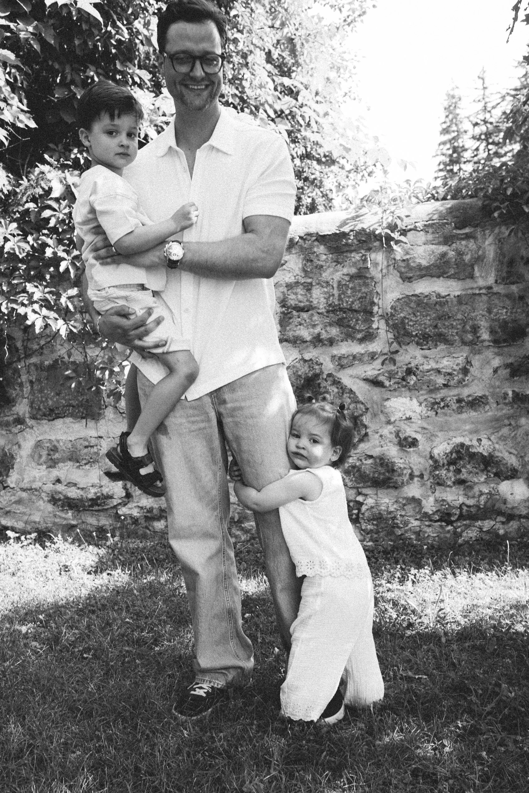 A man holding a young boy in his arms, standing outdoors with a stone wall and trees in the background. A young girl is hugging the man's leg, looking at the camera.