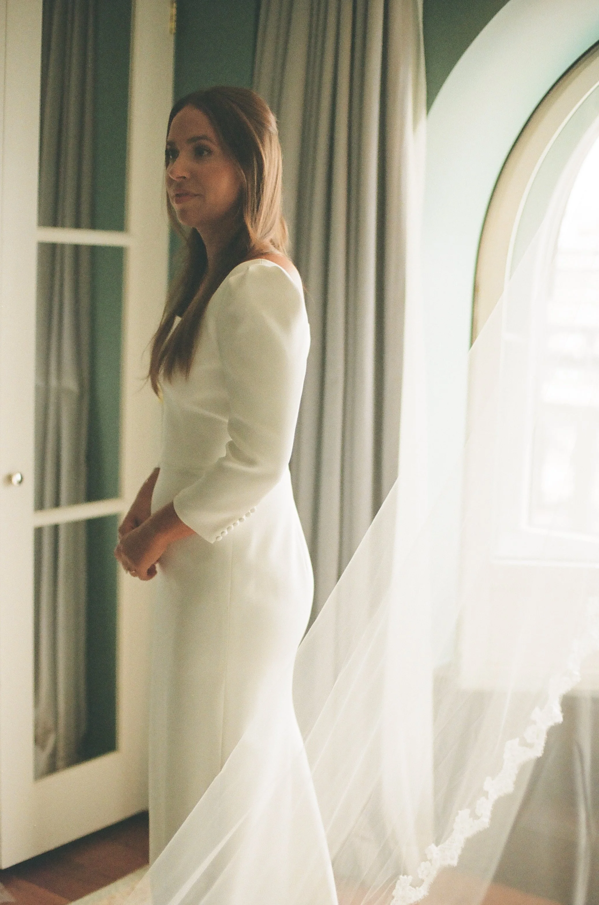 A woman in a white dress standing near a window with curtains, with sunlight illuminating her.