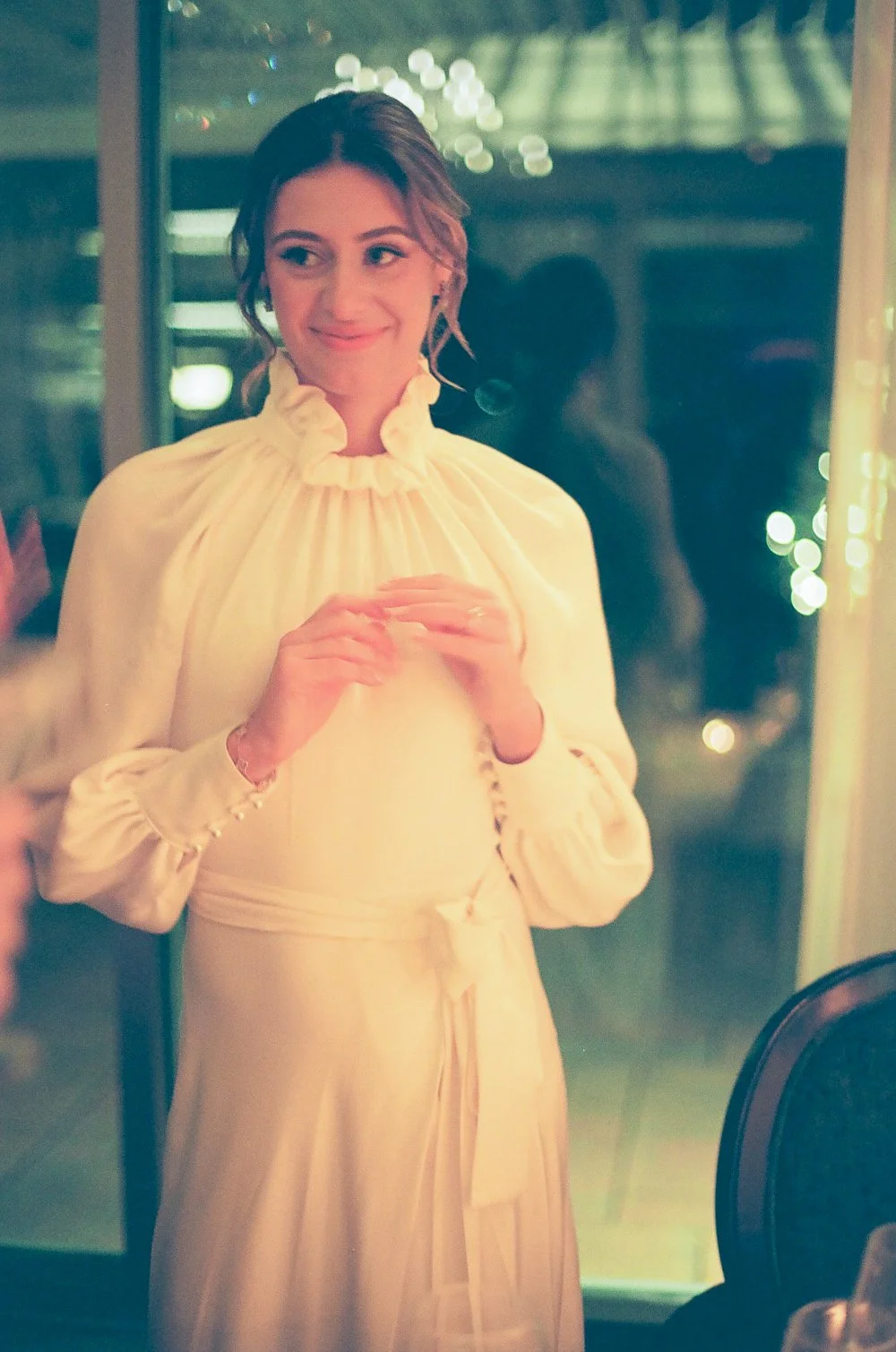 A woman in a vintage cream-colored dress with a high ruffled collar and long sleeves, standing indoors near a window with fairy lights in the background.