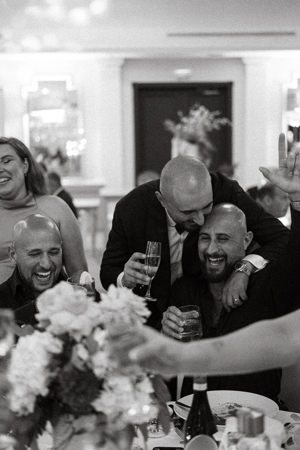 People celebrating at a formal event, with three men and one woman smiling and holding drinks, including champagne glasses, in a decorated indoor venue.
