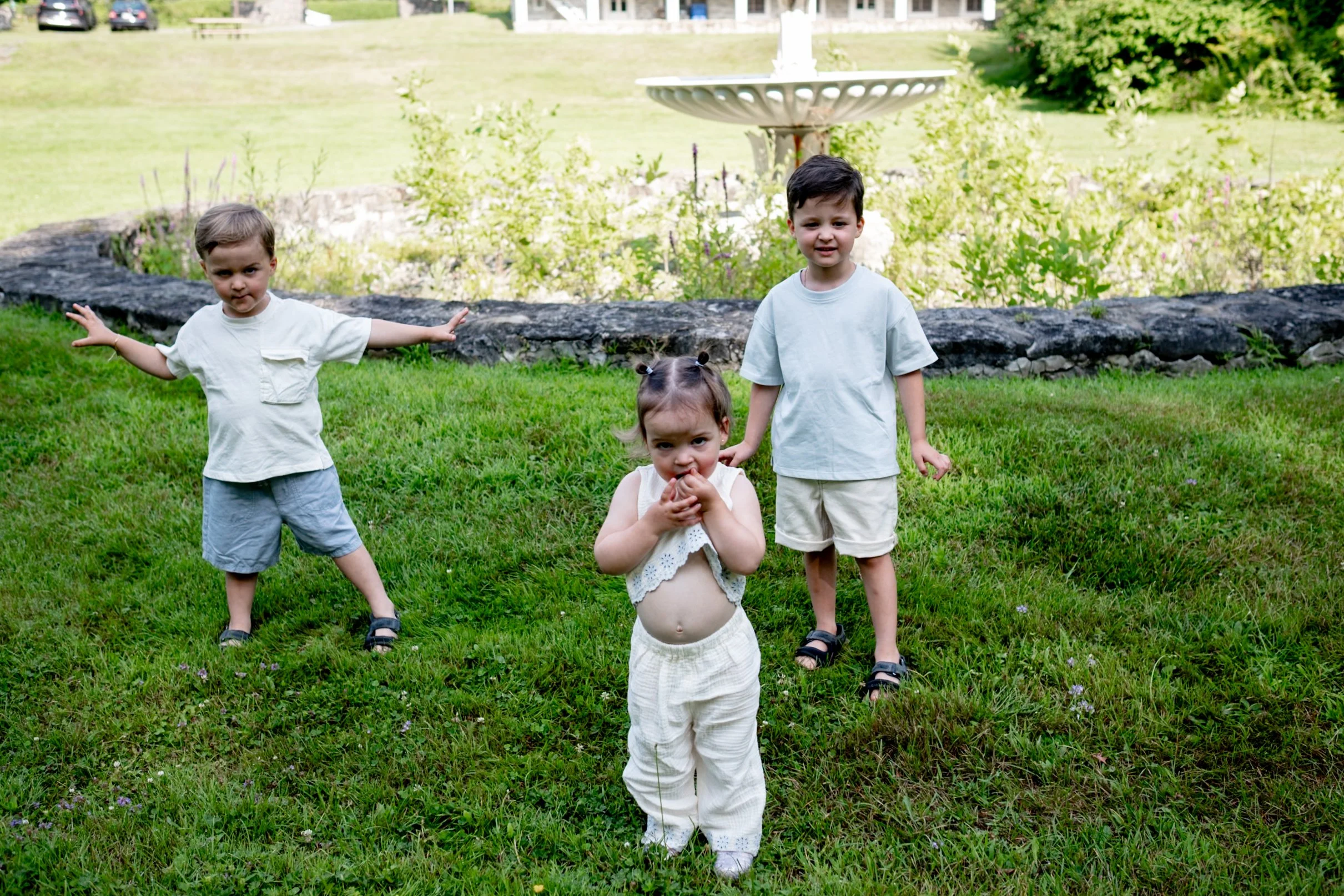 Three children and one toddler girl standing on grass in a backyard with a stone border and plants in the background, with a bird bath visible.