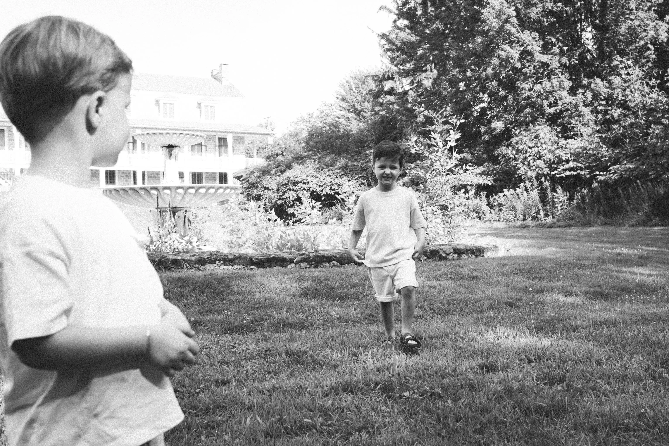 Two young boys playing outdoors near a fountain and trees, with a large house in the background, in a black-and-white photograph.