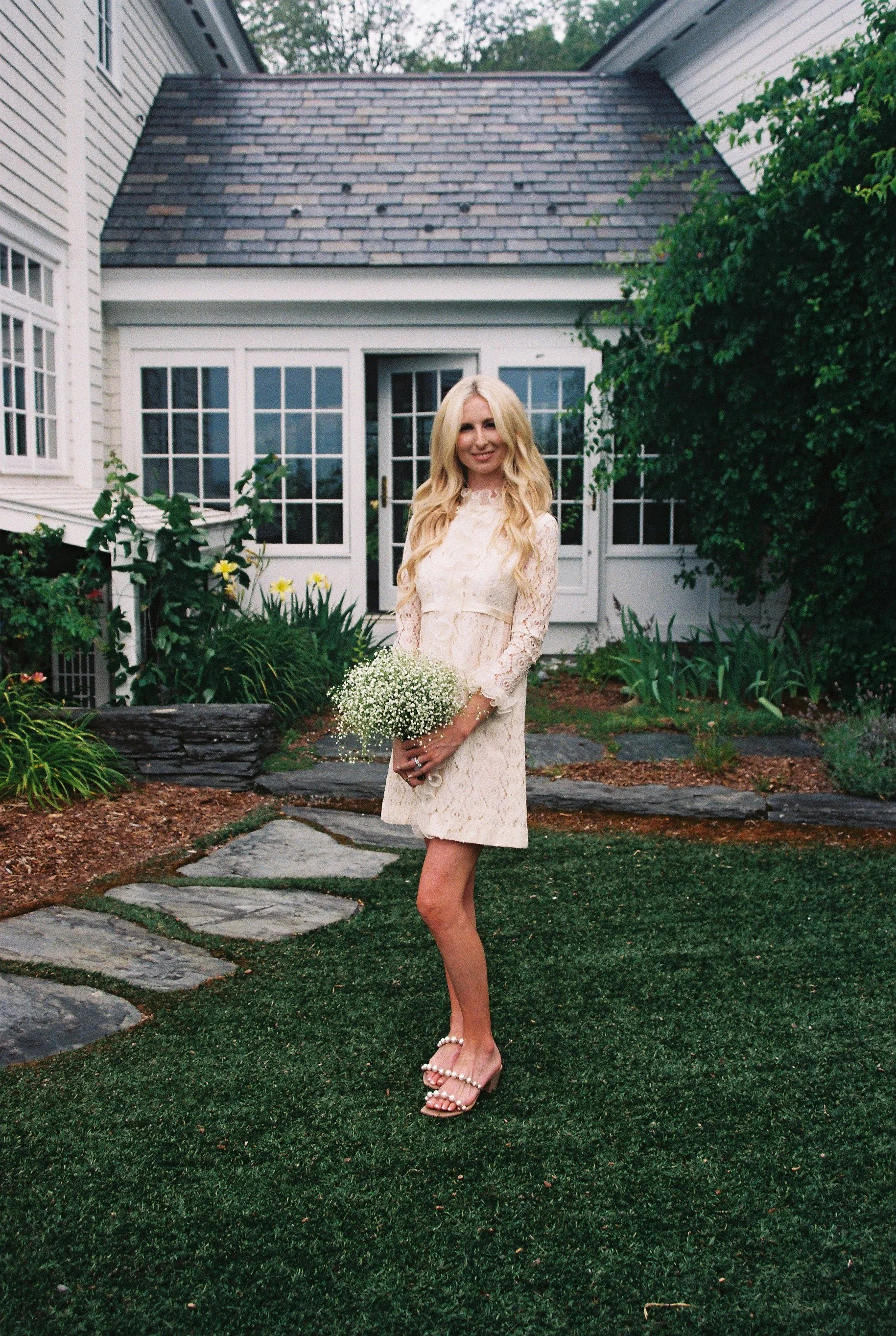 A woman with long blonde hair wearing a cream lace dress and pearl-embellished heels, holding a bouquet of white baby's breath flowers, standing on a stone pathway in a garden with green plants and a white house with large windows and a glass door in