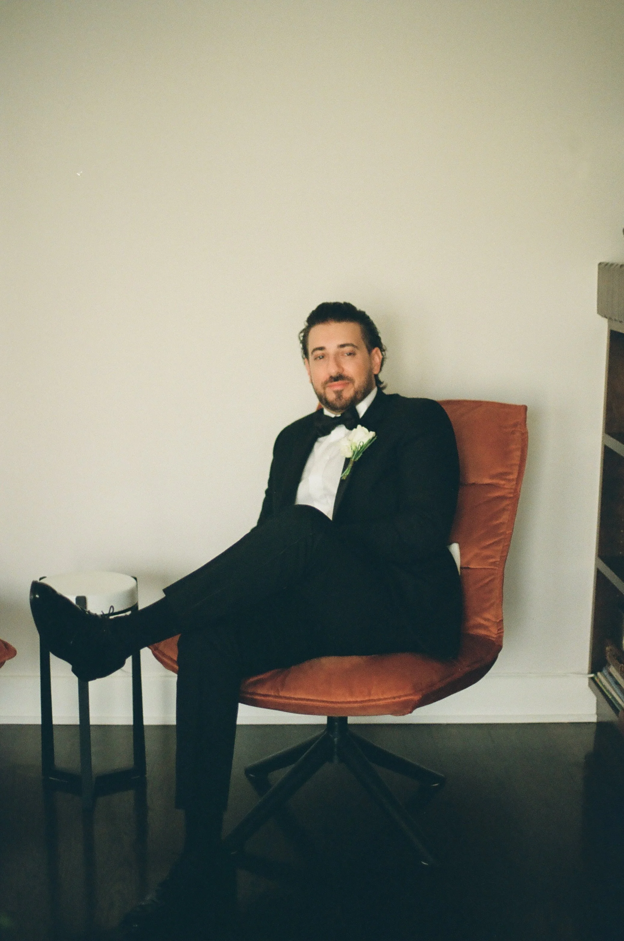 A man in a tuxedo sitting on an orange chair with his legs crossed. He has dark, wavy hair and a beard, and is wearing a white dress shirt, black bow tie, and a white boutonniere. A small table with a white hat or box is next to him, and a bookshelf 