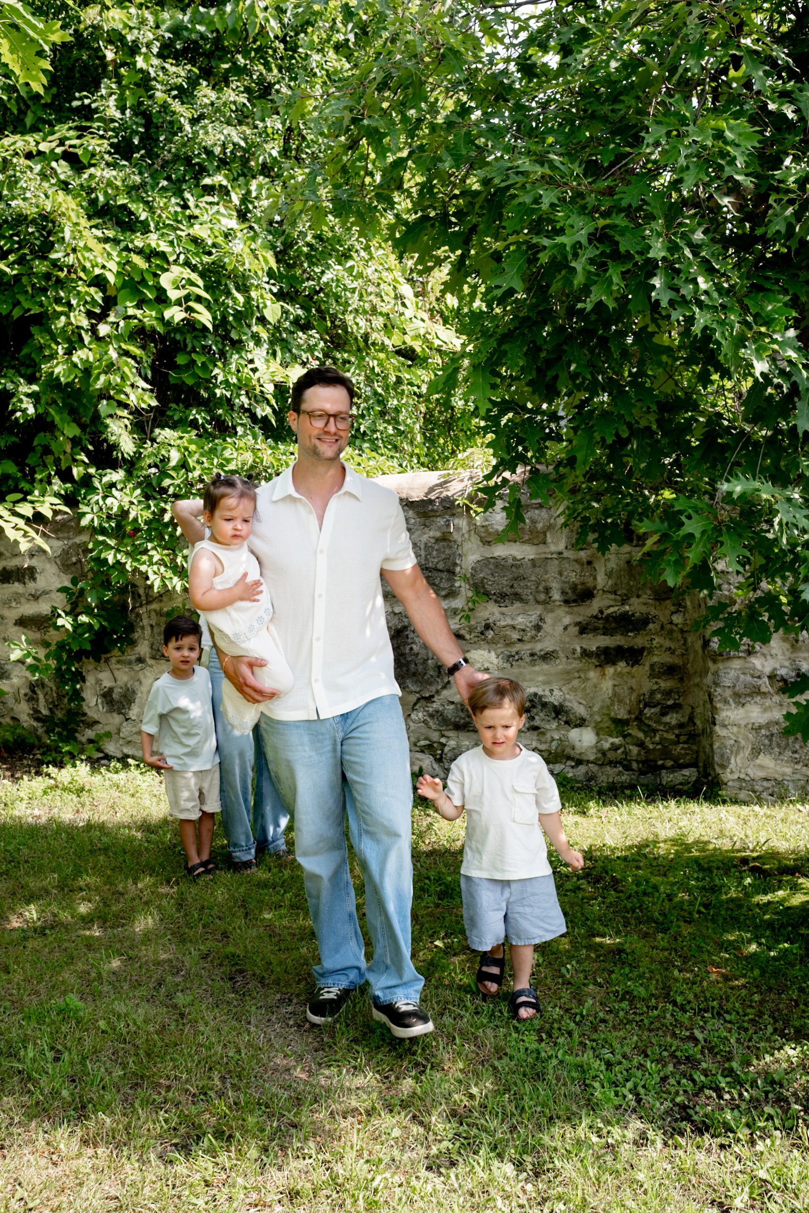 A man walking outdoors with four children, overgrown trees with lush green leaves in the background, and a stone wall behind them.