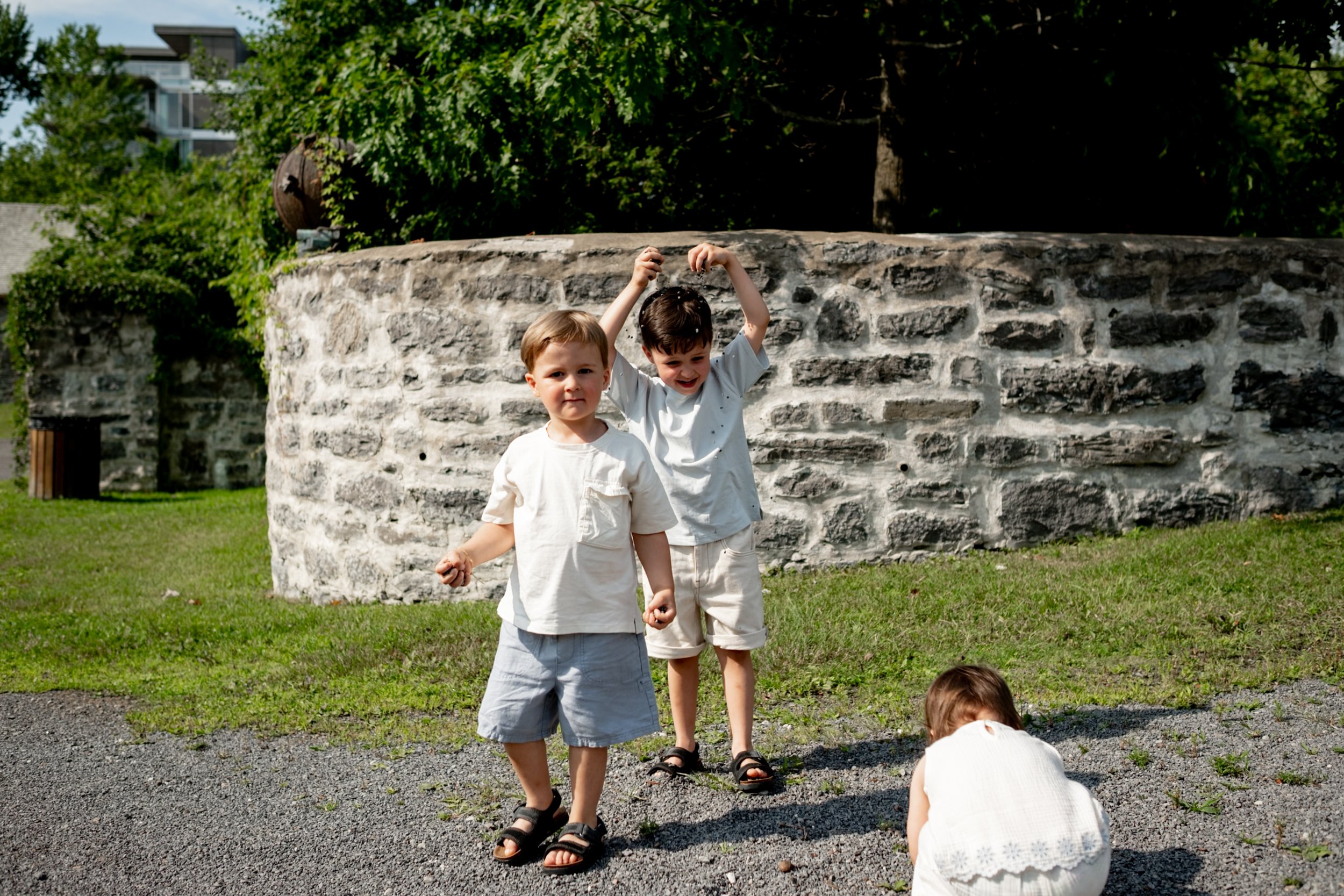 Two young boys standing outside near a stone wall with lush greenery and trees in the background. One boy is looking at the camera with a slight smile, while the other boy is smiling and raising his arms.
