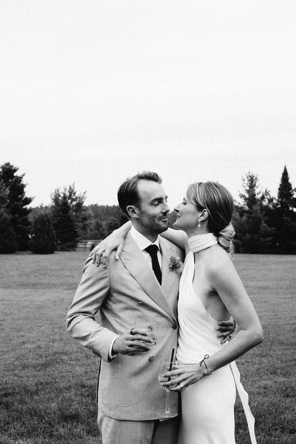 Black and white photo of a couple at their wedding, standing outdoors with trees in the background. The groom is in a light-colored suit holding a glass of drink, while the bride is in a sleeveless dress with a halter neck, leaning in for a close moment, both smiling.