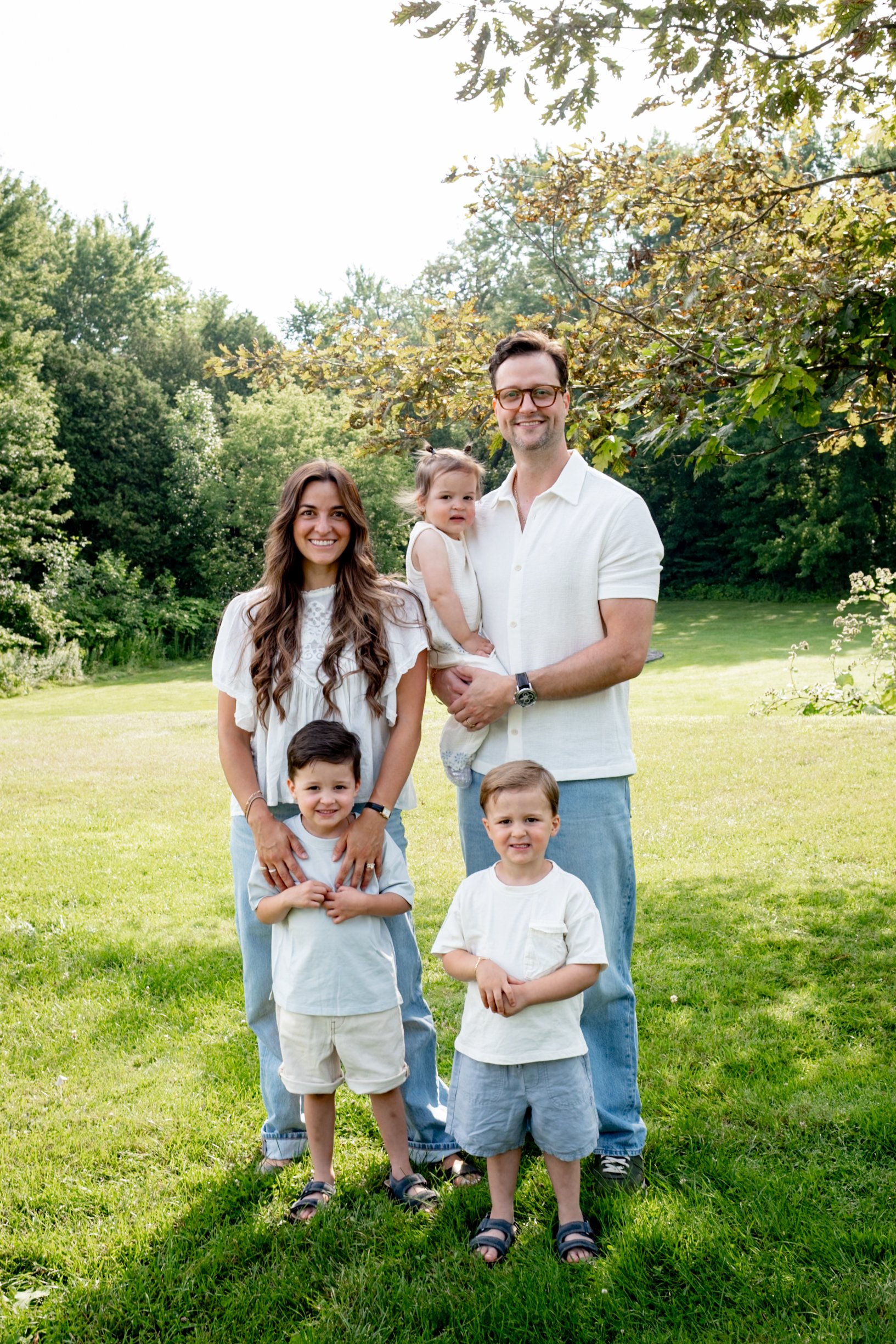 Family of seven standing outdoors on a sunny day, surrounded by green trees and grass. The family includes two adults and five children, all smiling and dressed casually in light-colored clothes.