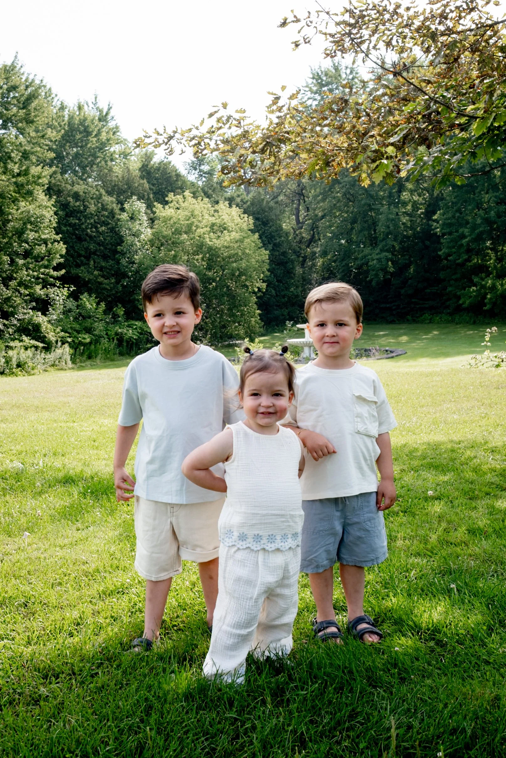 Group of four children standing in a grassy backyard, surrounded by trees, with a fountain in the background and bright sunlight overhead.