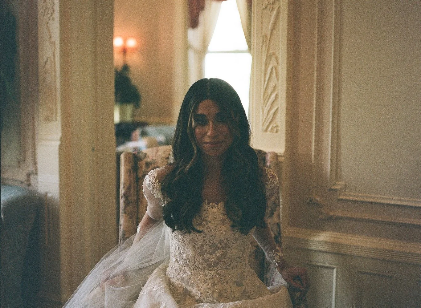 A woman in a wedding dress sitting in a vintage room with ornate wall paneling, a floral armchair, and window curtains, smiling softly.