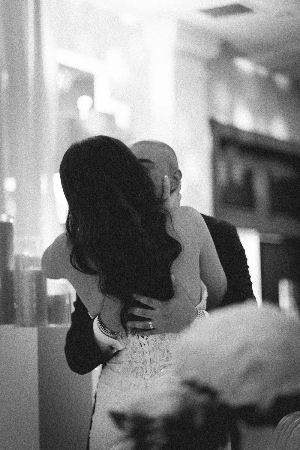 A couple sharing a kiss, with the woman in a wedding dress and the man in a suit, in a softly lit indoor setting.