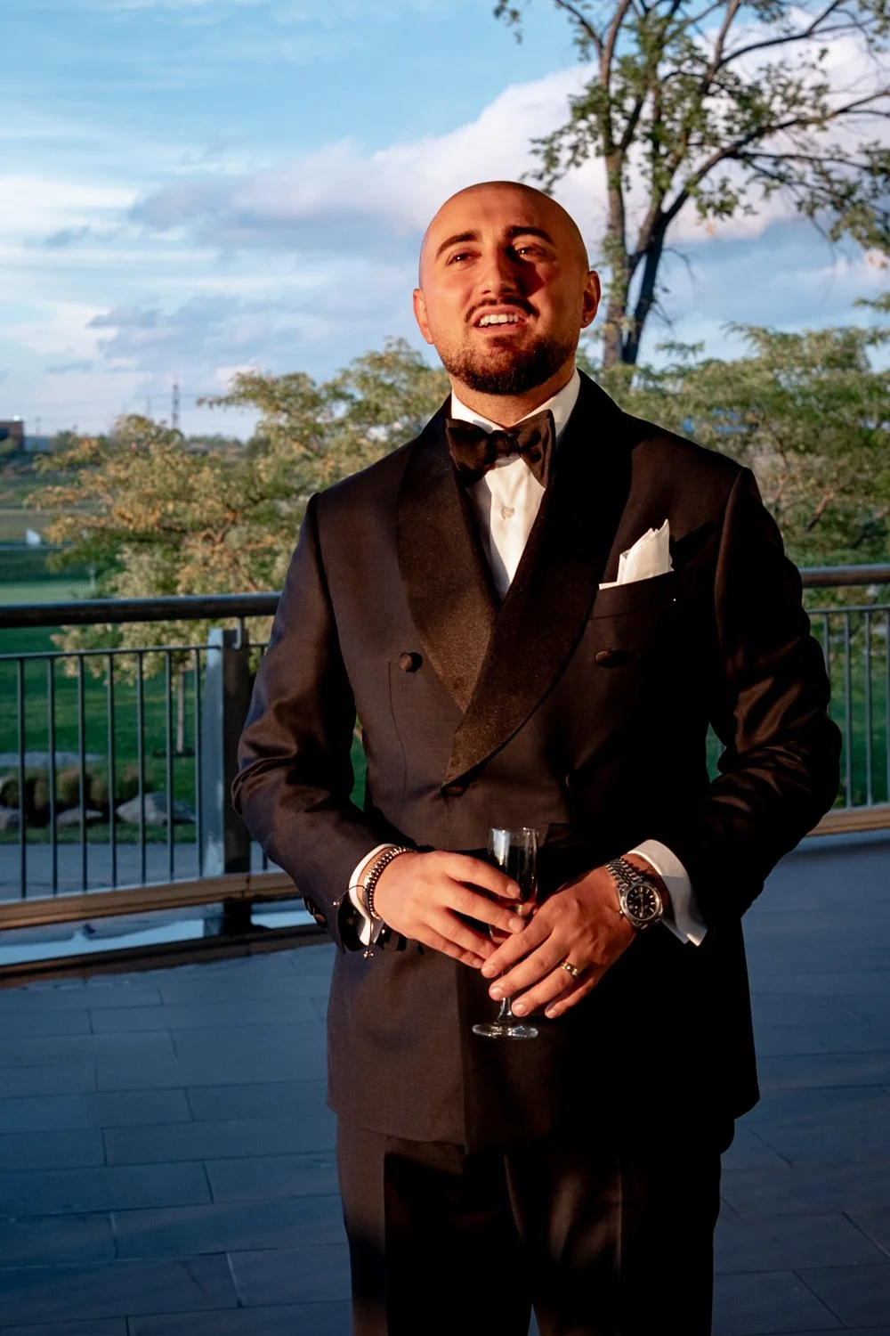 A groom in a tuxedo holding a glass of champagne on a balcony with trees and a partly cloudy sky in the background.
