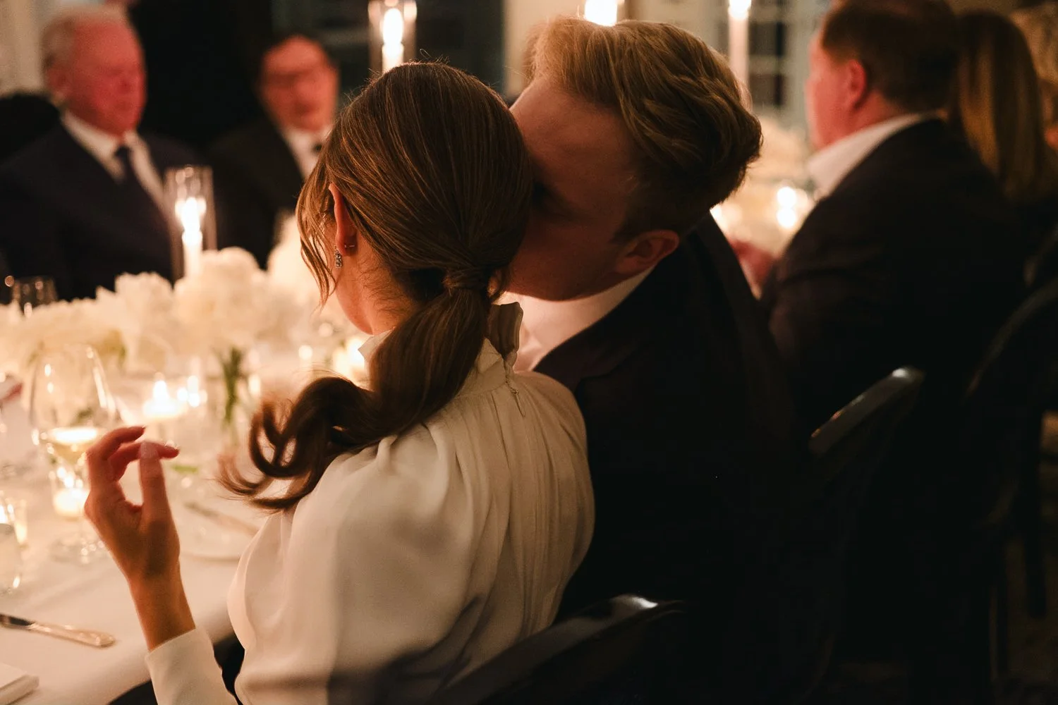A couple sharing a kiss at a formal dinner event, surrounded by other guests and candlelit decor.