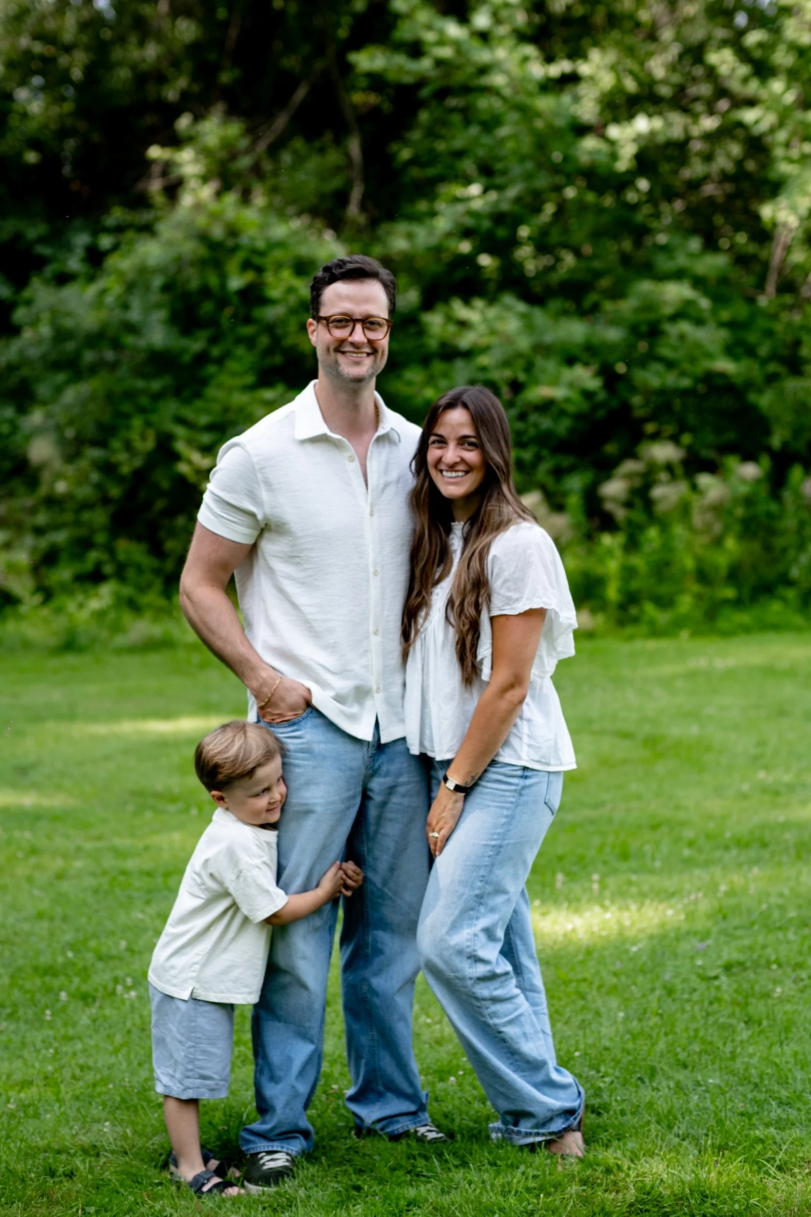 A family of four standing on grass in a park with lush green trees in the background. The grown man and woman are smiling at the camera, with the man wearing glasses, a white shirt, and light blue jeans, and the woman wearing a white blouse and light