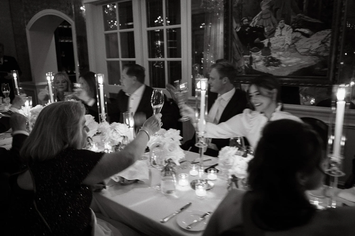 People celebrating at a formal dinner party, raising champagne glasses, in a decorated dining room with candles and artwork on the wall.