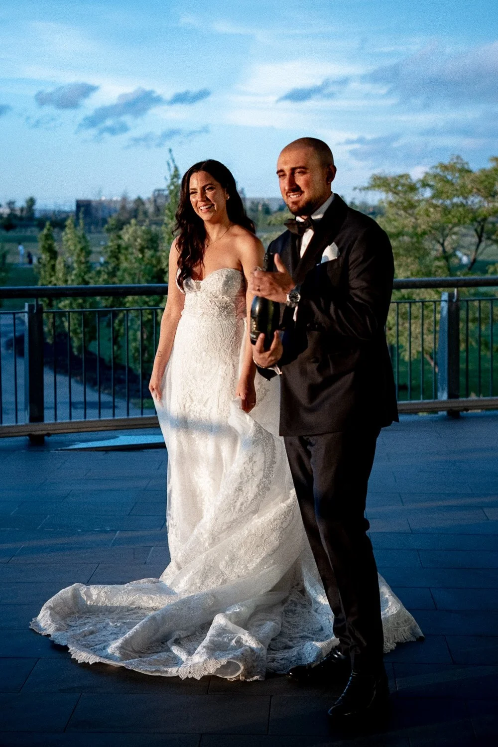 A bride in a white wedding dress and a groom in a black tuxedo stand on a balcony, with a cityscape and trees in the background, during a wedding celebration at sunset.