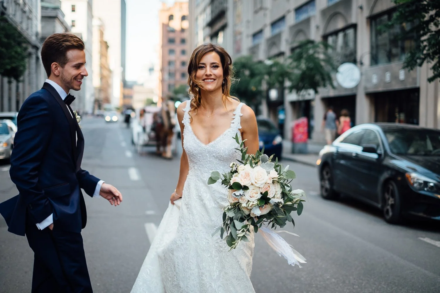 A bride in a white wedding dress holding a bouquet of white and pink roses, smiling on a city street, with a groom in a dark suit standing nearby.