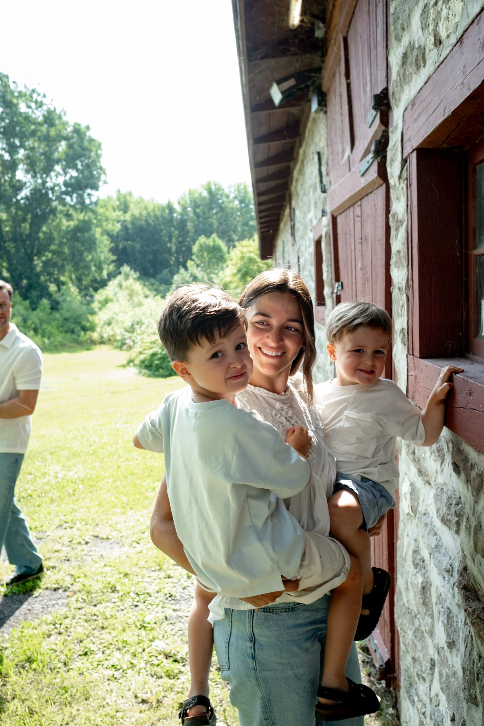 A woman carrying two young children on her hips outside a stone and wood building, smiling. An adult man stands in the background on a sunny day with trees.