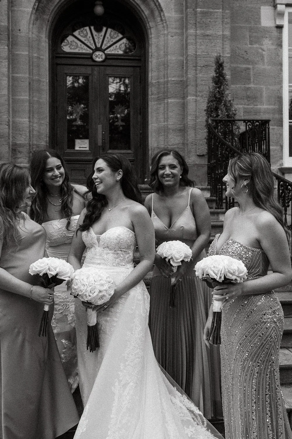 Black and white photo of six women in formal dresses, one in a wedding gown, standing on steps outside a building, holding bouquets, smiling and engaging in conversation.