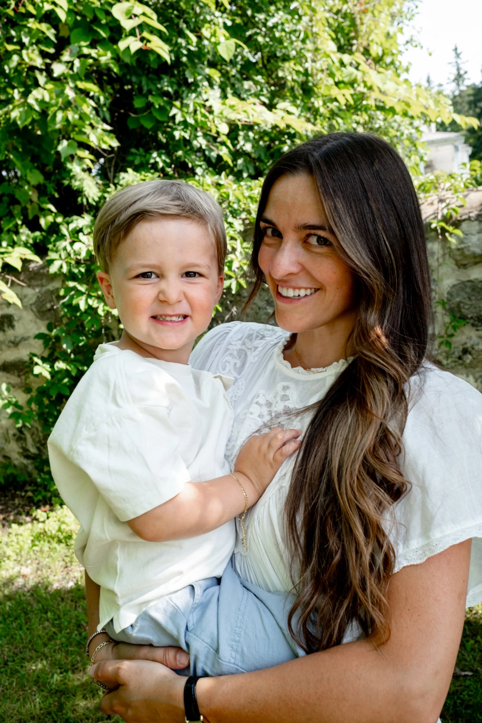 A woman with long dark hair holding a young boy with blonde hair outdoors in front of leafy green trees, both smiling.
