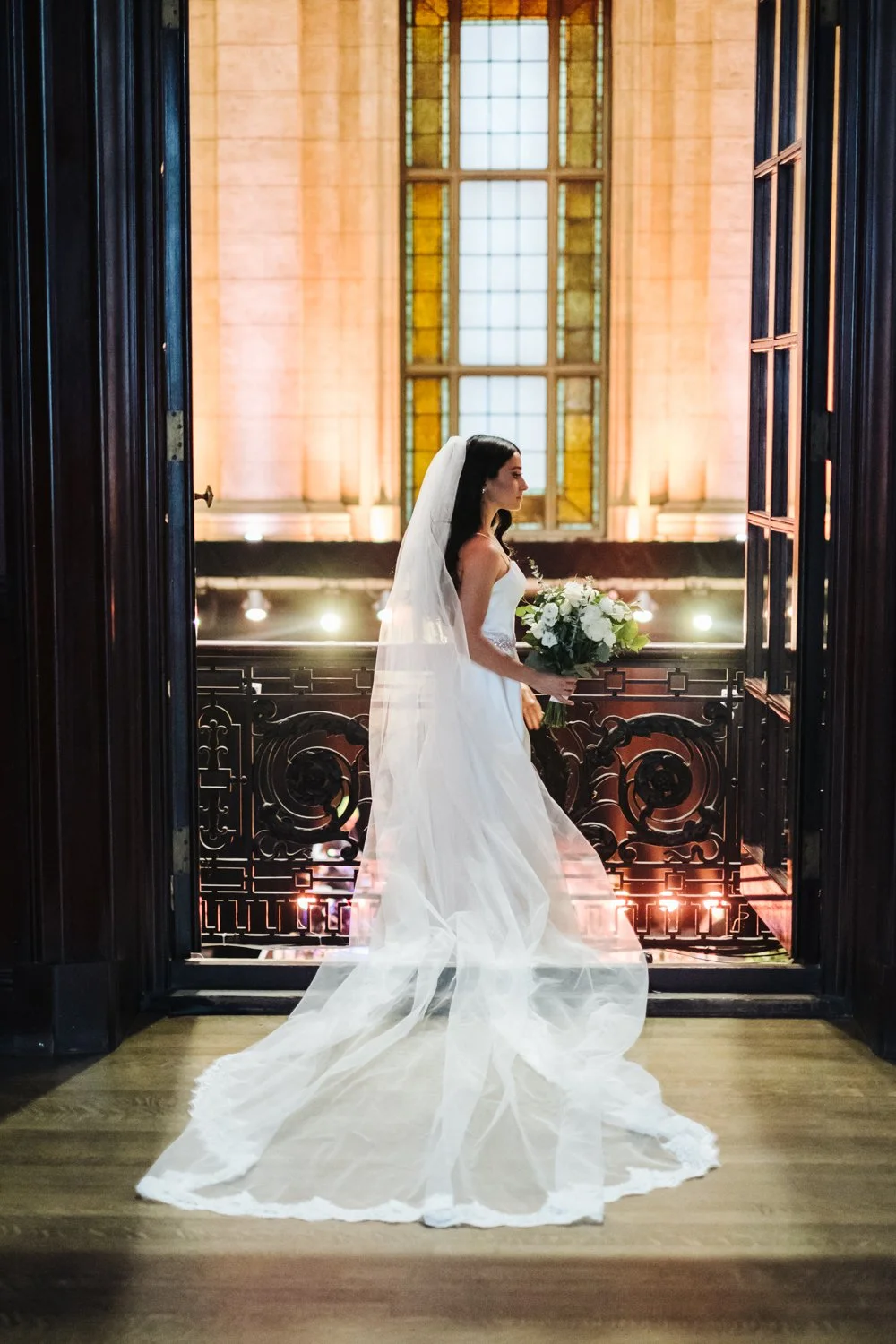 A bride in a white wedding dress and veil holding a bouquet of white flowers, standing on a wooden floor in front of an ornate iron railing and large stained glass window, lit with warm lighting.