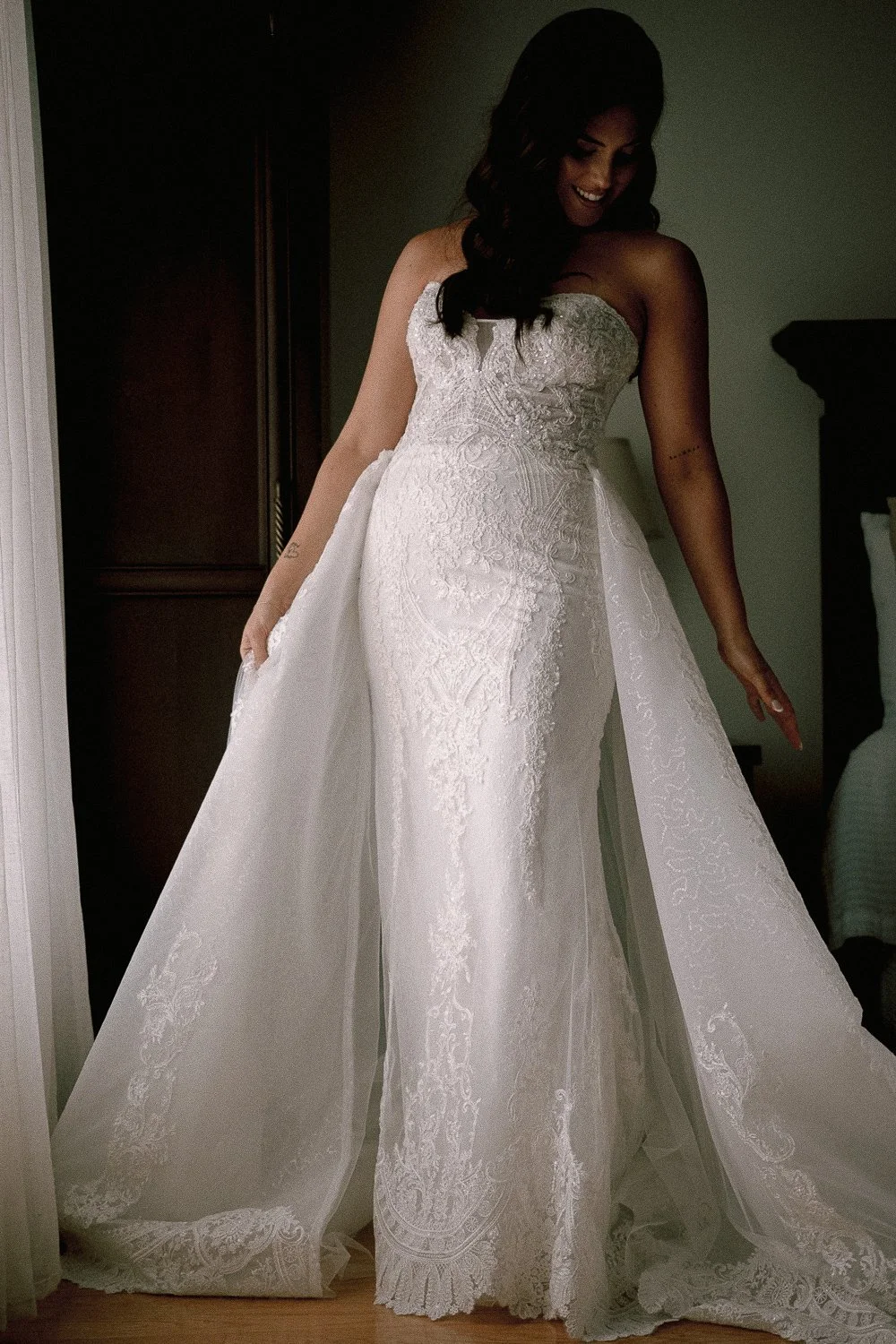 Bride in a wedding dress, standing in a room, looking down, holding the train of her dress.