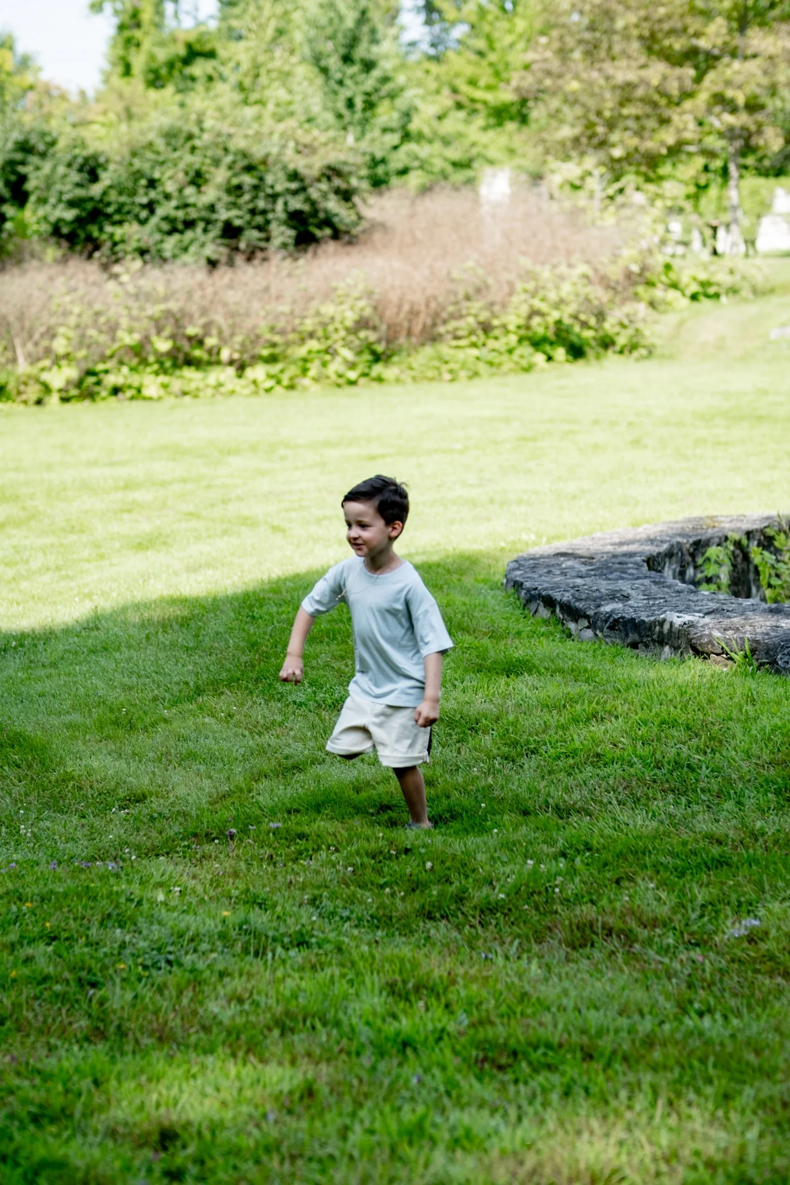 A young boy playing outside on a grassy lawn, smiling and wearing a light-colored t-shirt and beige shorts, with a background of trees and bushes.