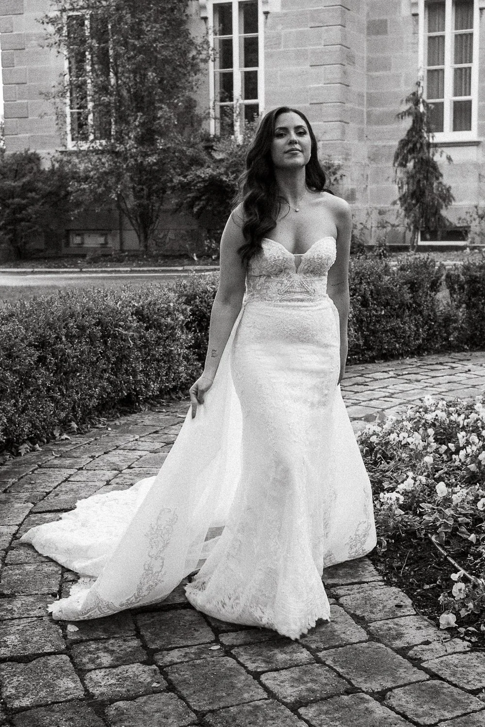 Montreal wedding photographer photographed a woman in a wedding dress standing on a cobblestone path with plants and a building in the background.