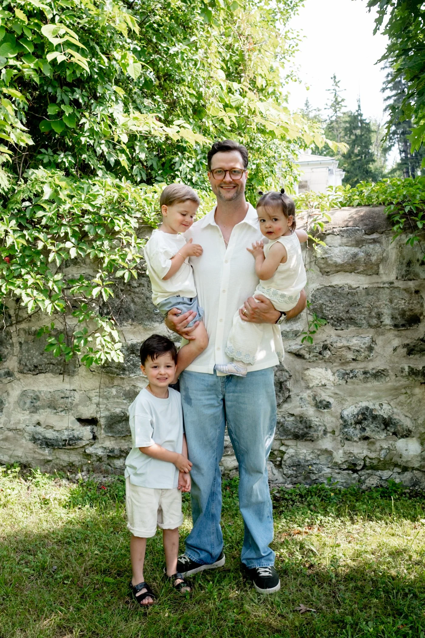 A man holding two young children outdoors in front of a stone wall with green foliage. Two other children stand on the grass at his feet.