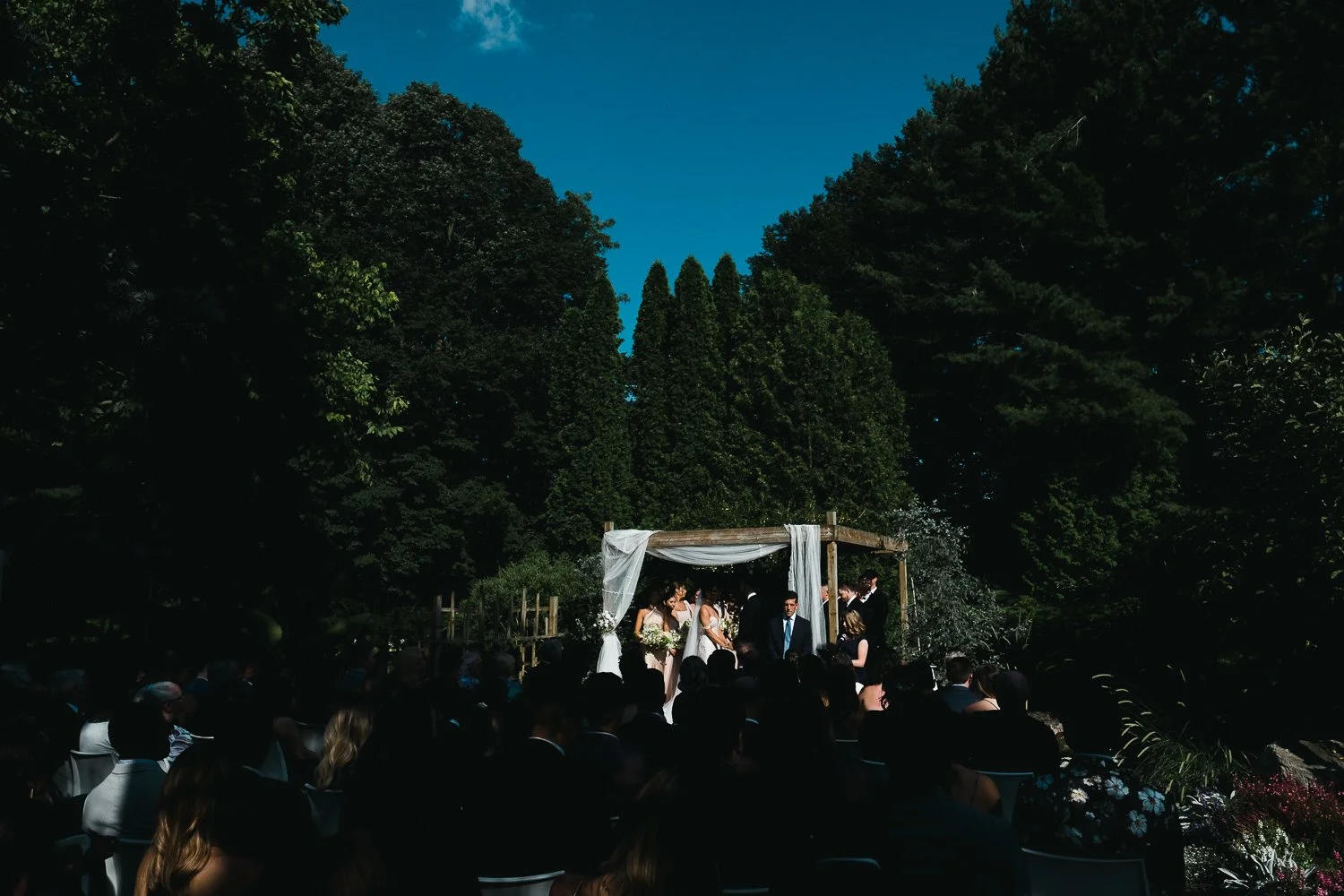 Outdoor wedding ceremony under a wooden arbor with sheer white fabric, set in a lush green garden with tall trees and a blue sky.