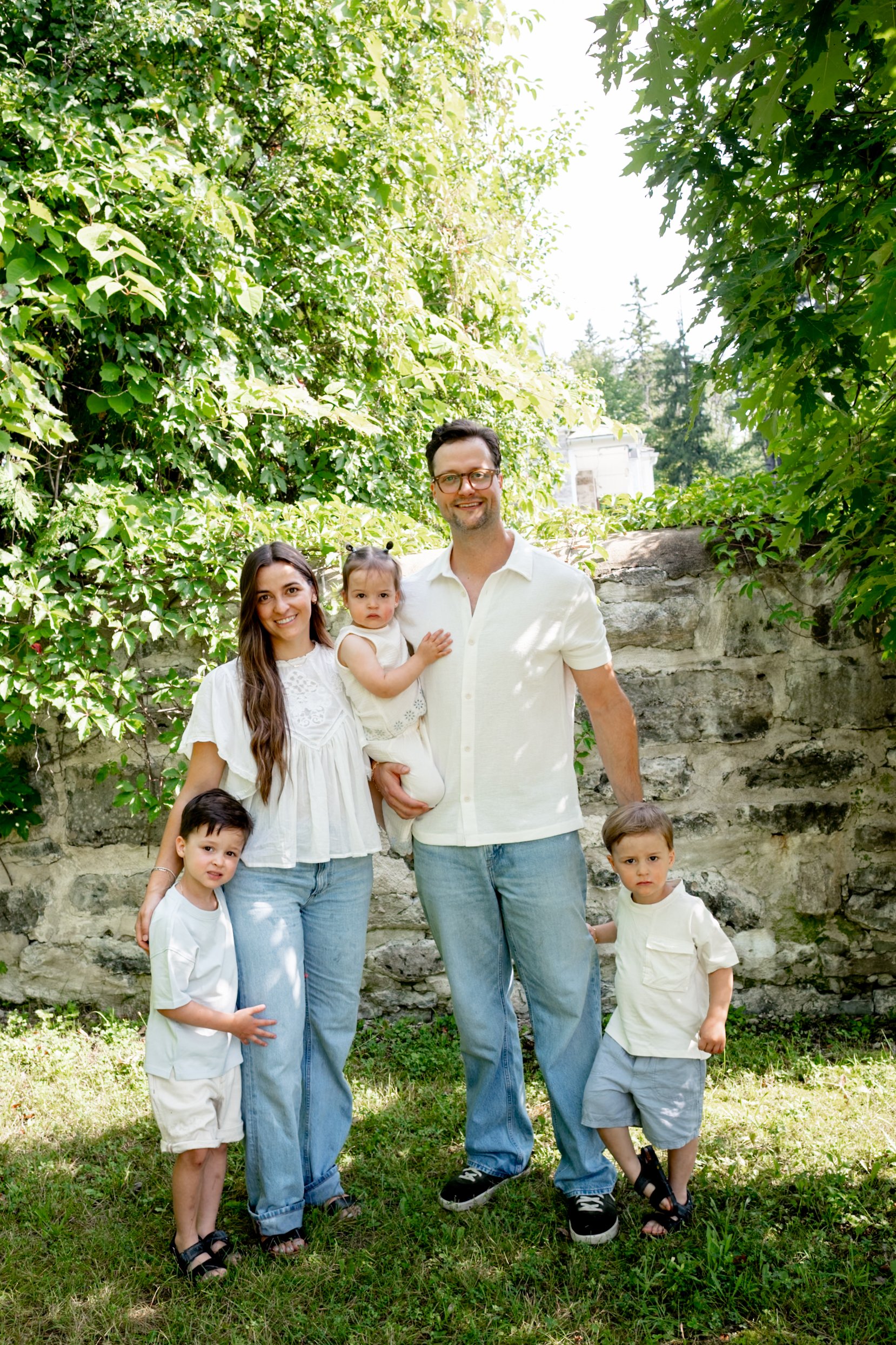 A family of seven standing outdoors in front of a stone wall and green foliage. The family includes two adult women, one adult man, and four children. They are dressed in light-colored clothing and appear to be enjoying a sunny day.
