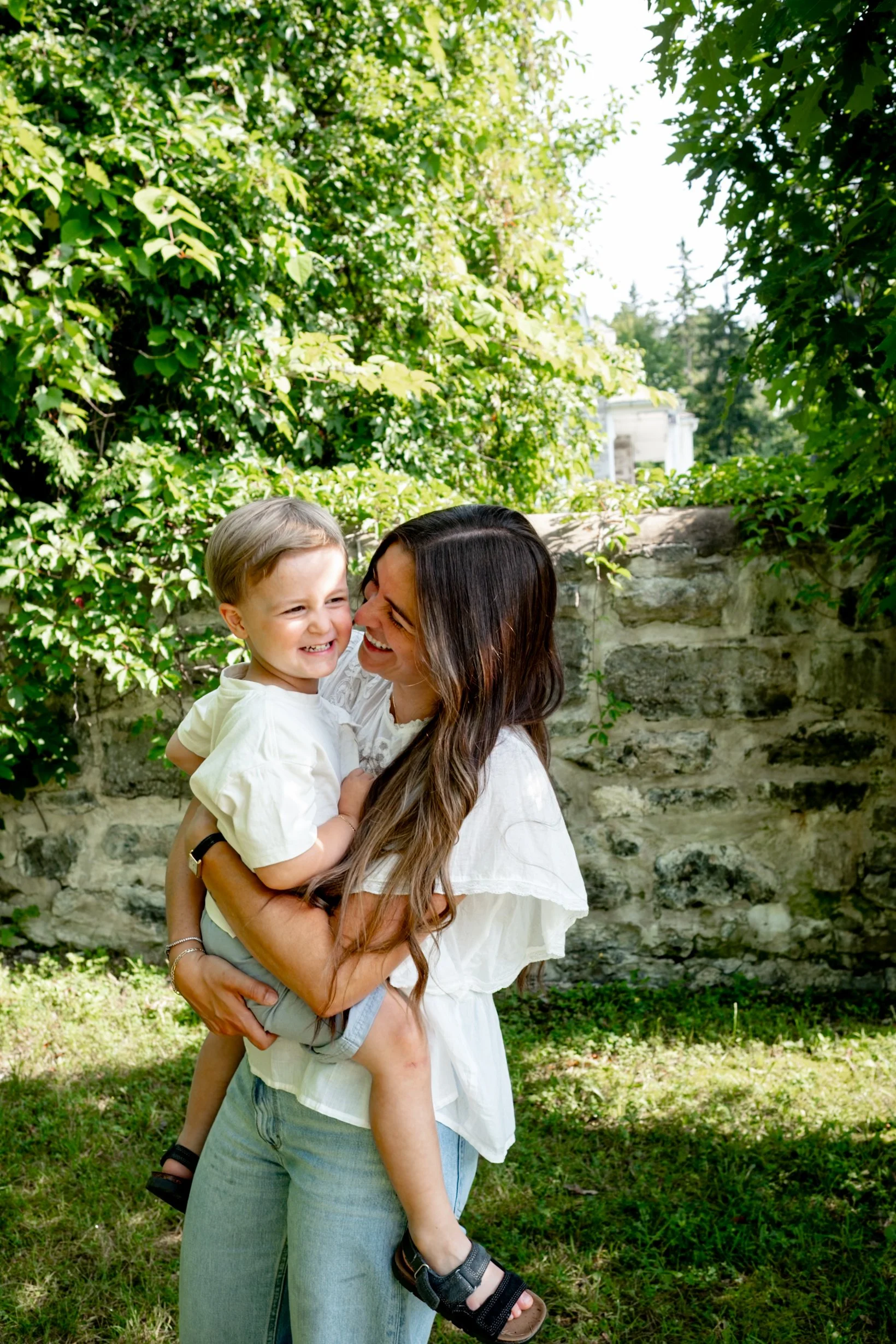 A woman holding a young boy outdoors surrounded by green trees and foliage, with a stone wall and a building in the background, both smiling and enjoying each other's company.