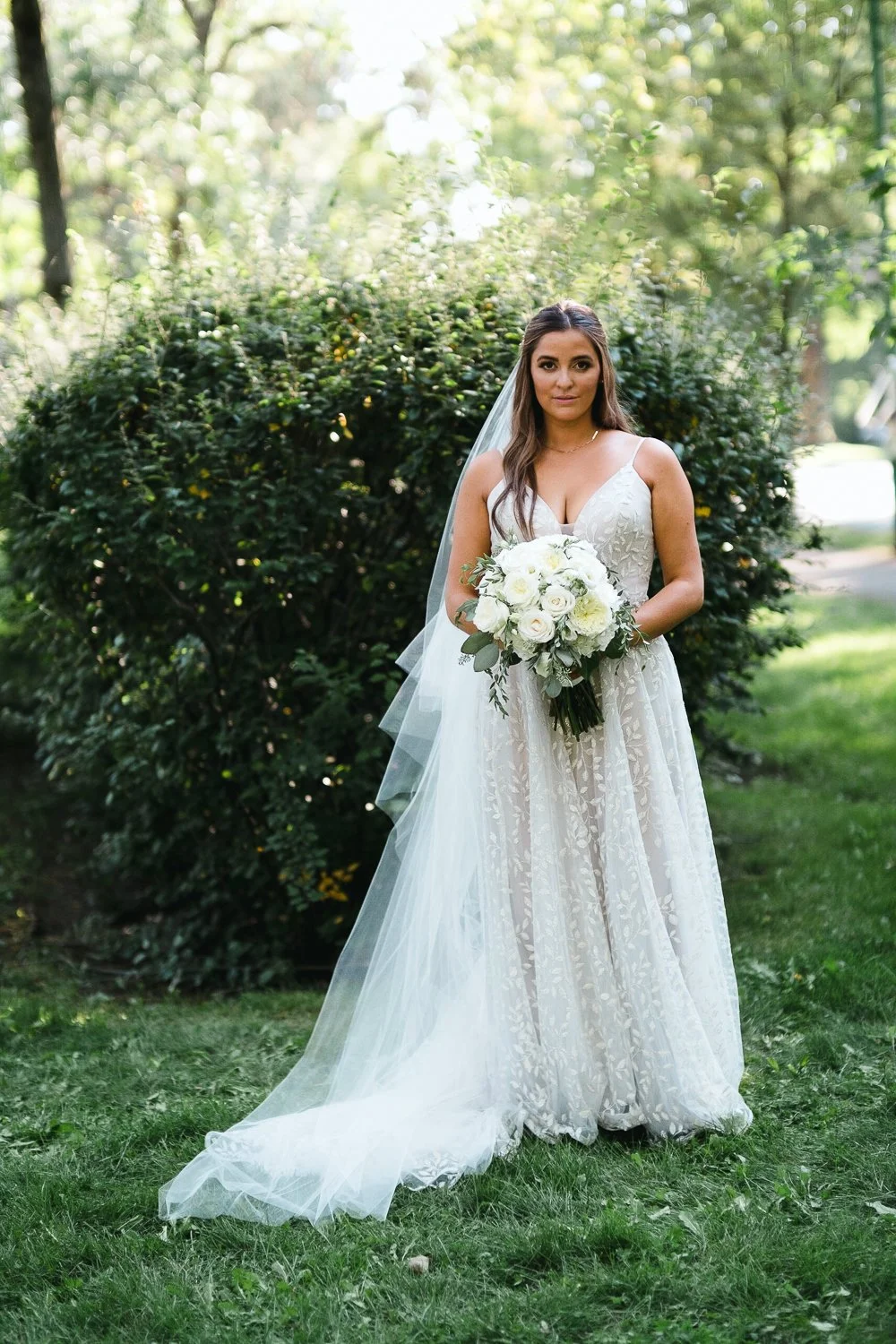 A bride standing outdoors on grass, wearing a white wedding dress and veil, holding a bouquet of white roses and greenery, with a background of green bushes and trees.