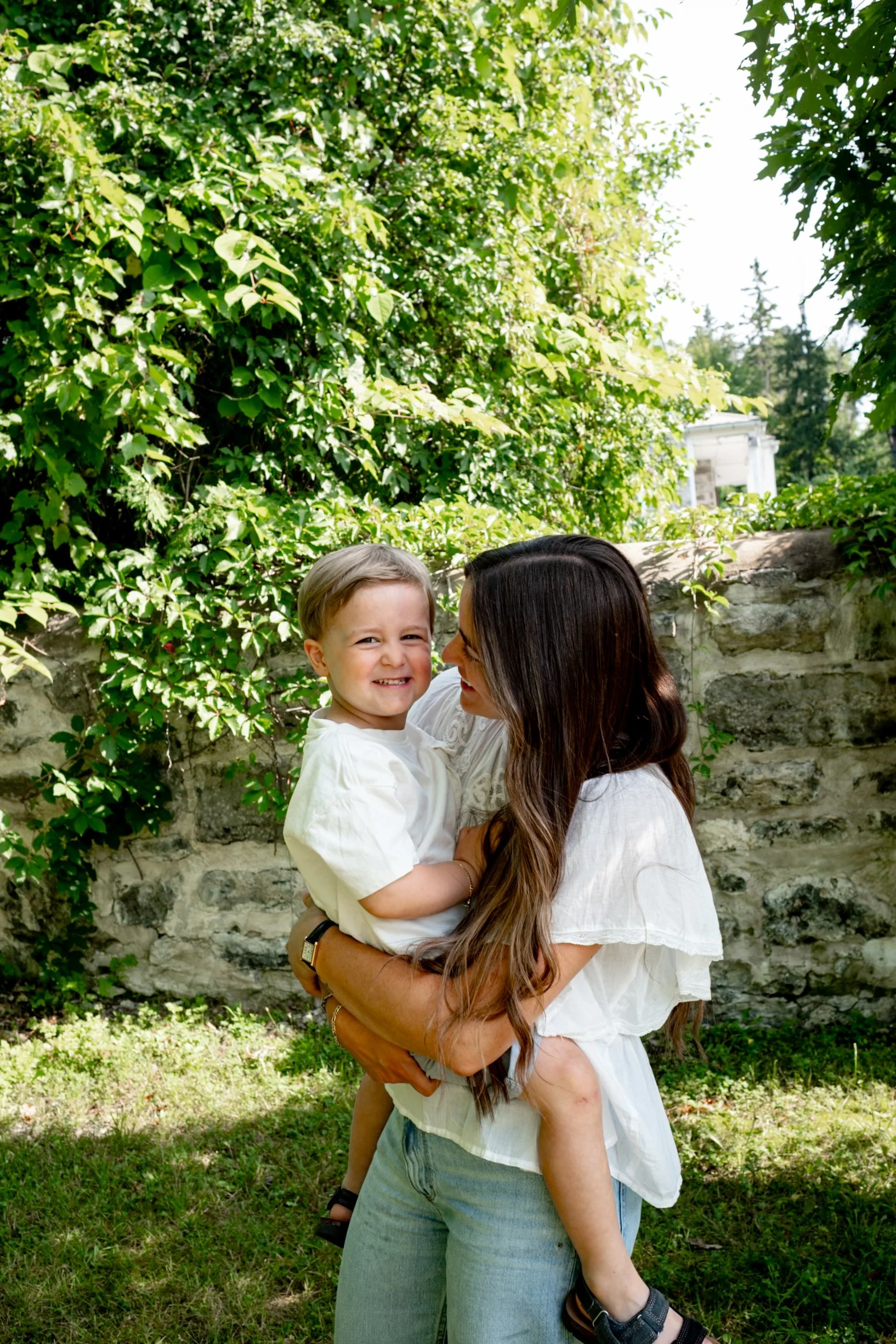 A woman with long brown hair is holding a smiling young boy in her arms. They are outdoors in front of a stone wall and surrounded by green foliage and trees.