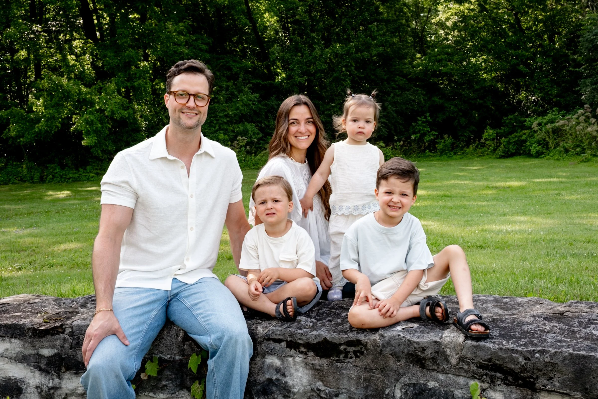 A family of six sitting on a log in a park with green trees in the background. The group includes two adults and four children. The adults are smiling, and the children have various facial expressions.