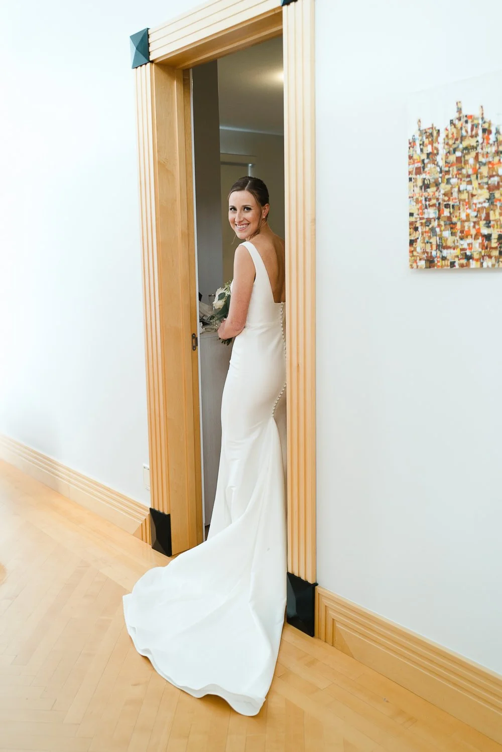 A smiling bride in a white wedding gown standing in a doorway, holding a bouquet of flowers, with a colorful abstract painting on the wall to her right.