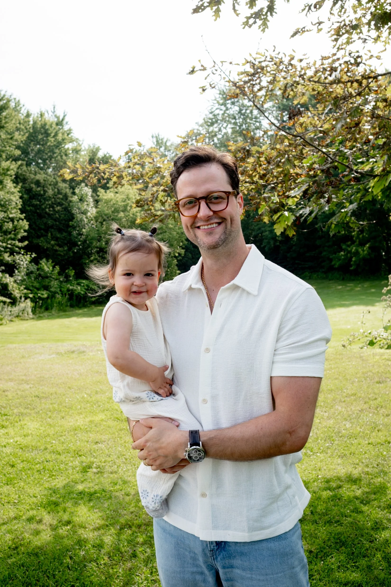 A man with glasses and a beard smiling while holding a young girl with pigtails outdoors in a park with lush green trees and grass.